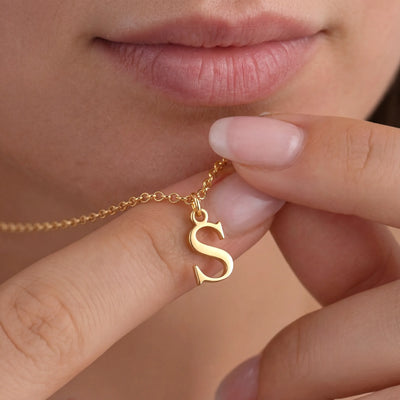 A close-up lifestyle shot of a person holding a delicate gold chain necklace with a polished gold initial 'S' pendant against their lips.